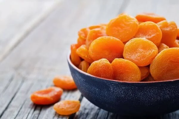 A dark bowl heaped with golden dried apricot halves on a weathered wooden table